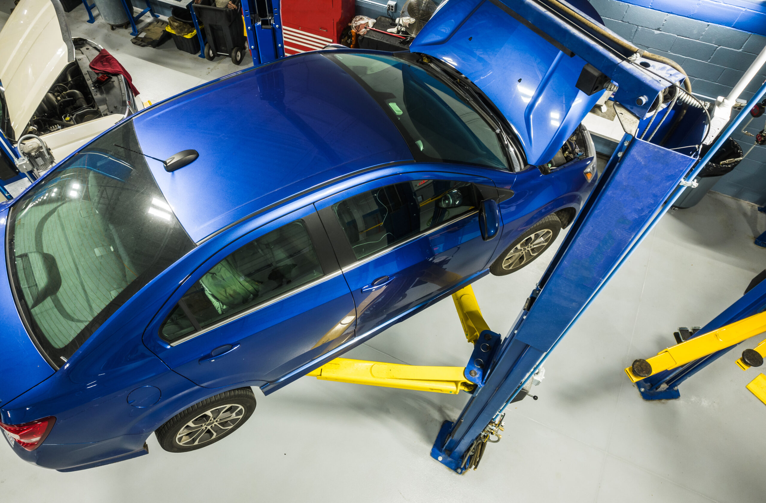 A modern compact car raised on a lift in a brightly lit service bay, with a mechanic inspecting its undercarriage.