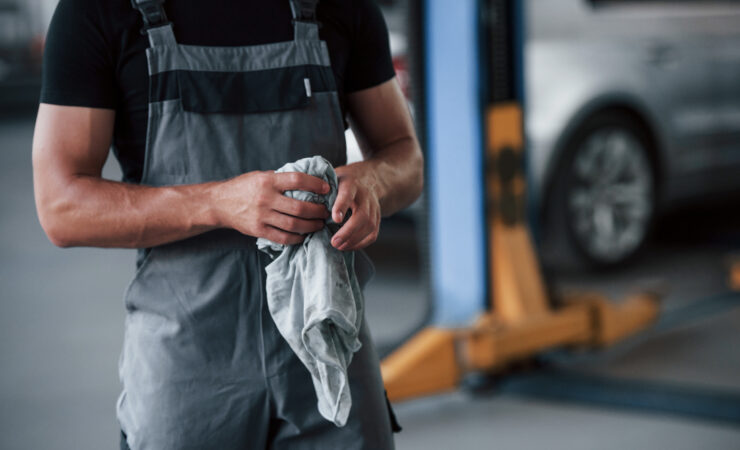 A man in a black shirt and grey uniform, with a satisfied expression, stands in a garage next to a repaired car.