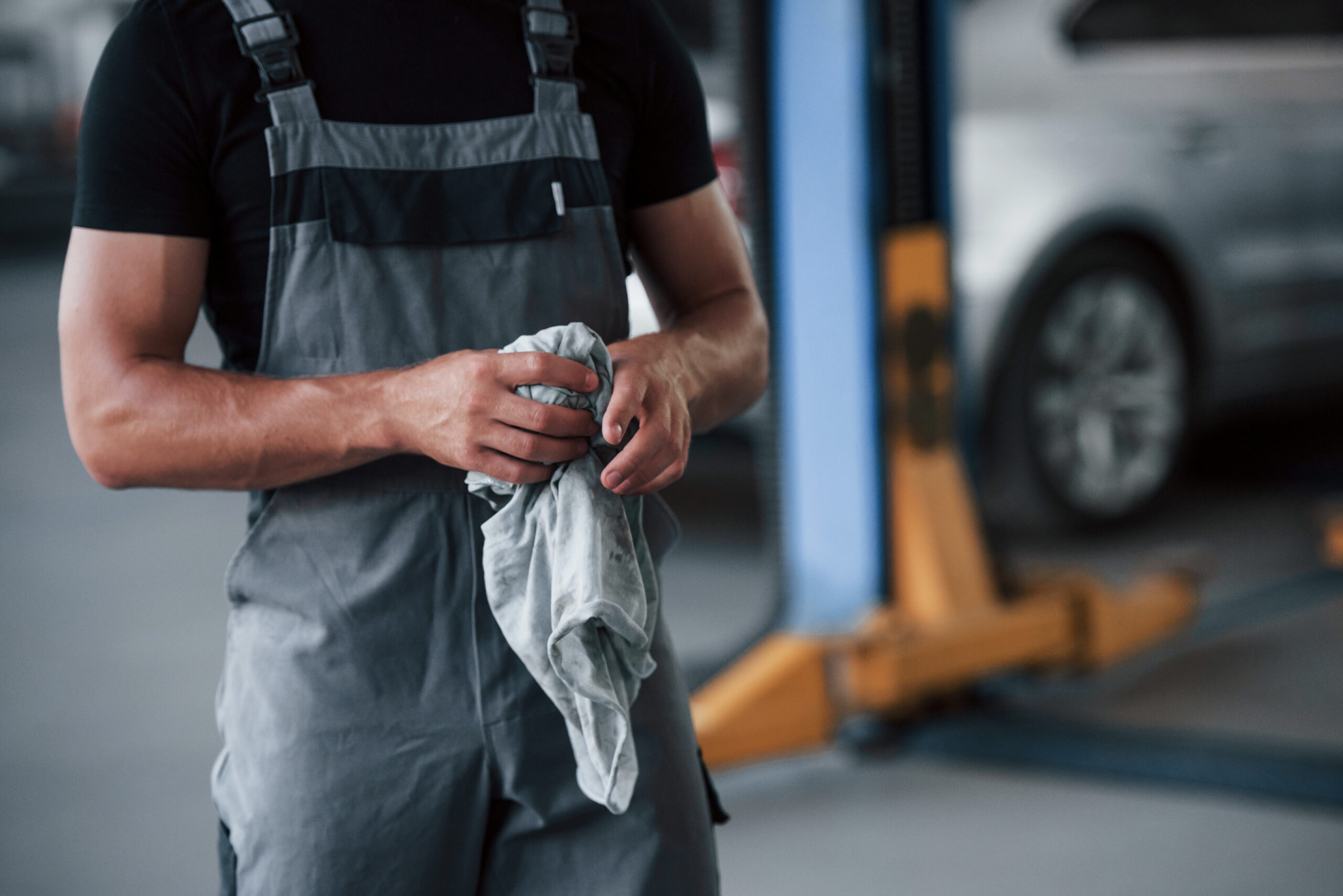 A man in a black shirt and grey uniform, with a satisfied expression, stands in a garage next to a repaired car.