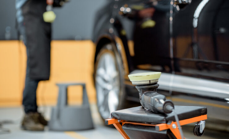 A professional car detailer uses a dual-action polisher with a wool pad to buff out scratches on a car's paint.