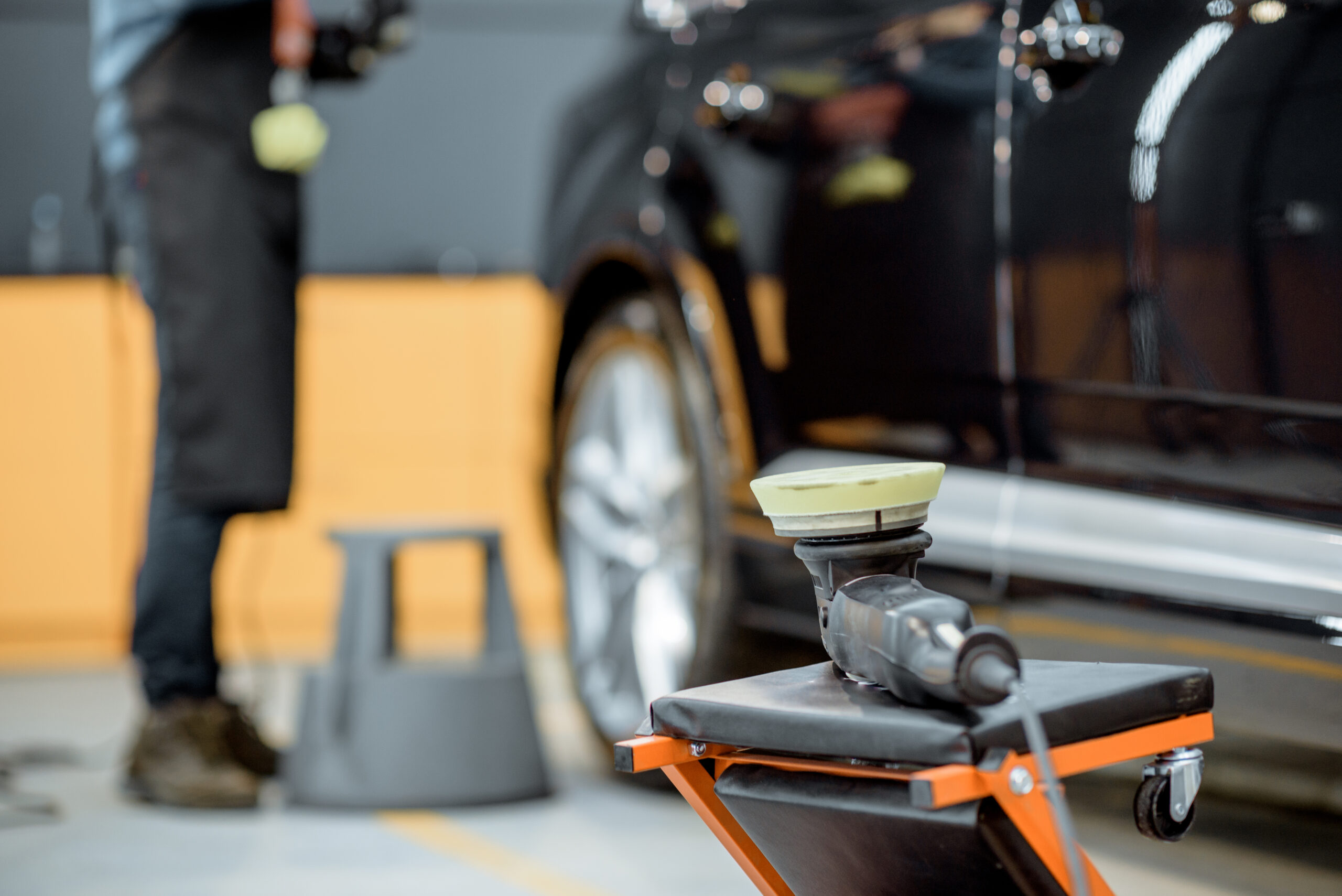 A professional car detailer uses a dual-action polisher with a wool pad to buff out scratches on a car's paint.