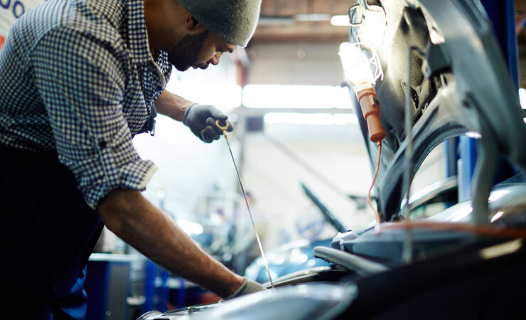 A certified mechanic, wearing a badge and uniform, smiles confidently while holding a diagnostic tool in front of a car.