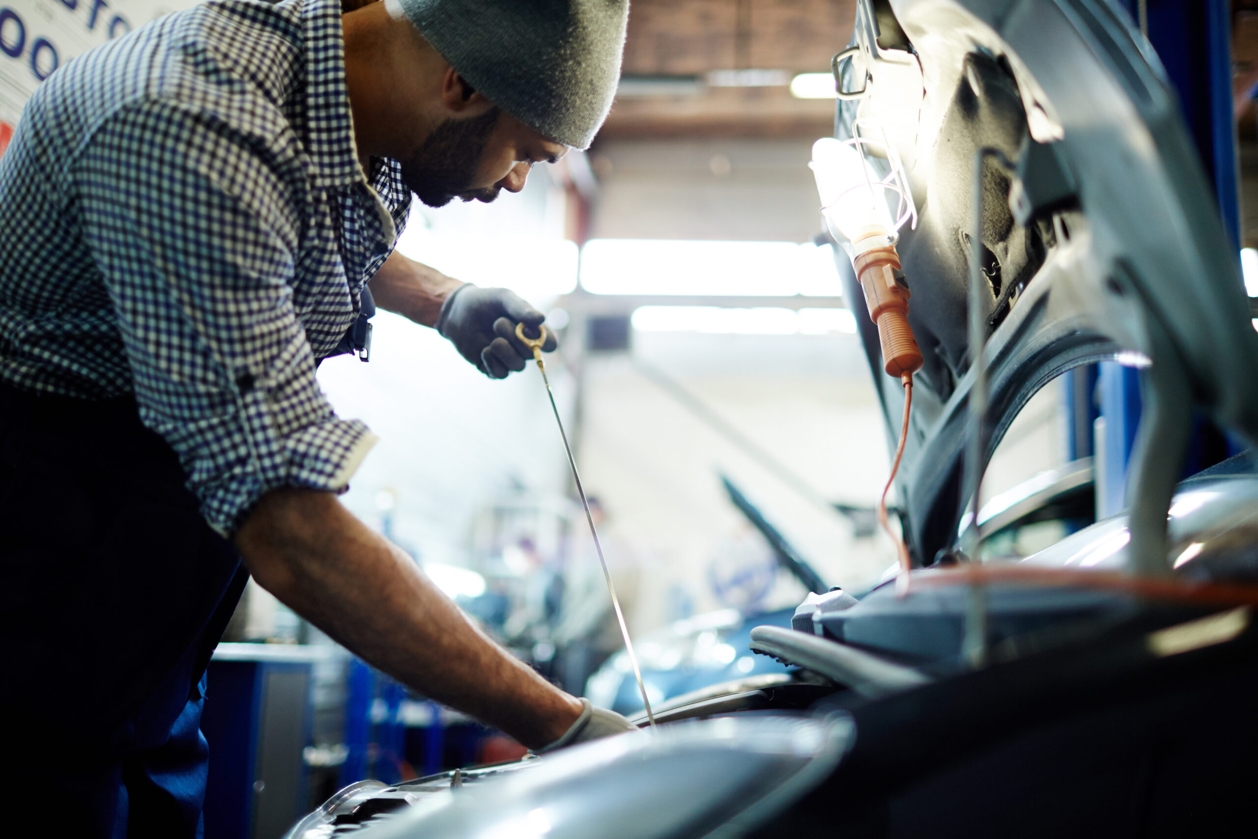 A certified mechanic, wearing a badge and uniform, smiles confidently while holding a diagnostic tool in front of a car.