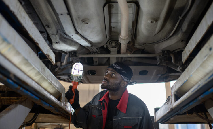 A focused young male auto technician, illuminated by a work lamp, inspects the undercarriage of a car during a repair.