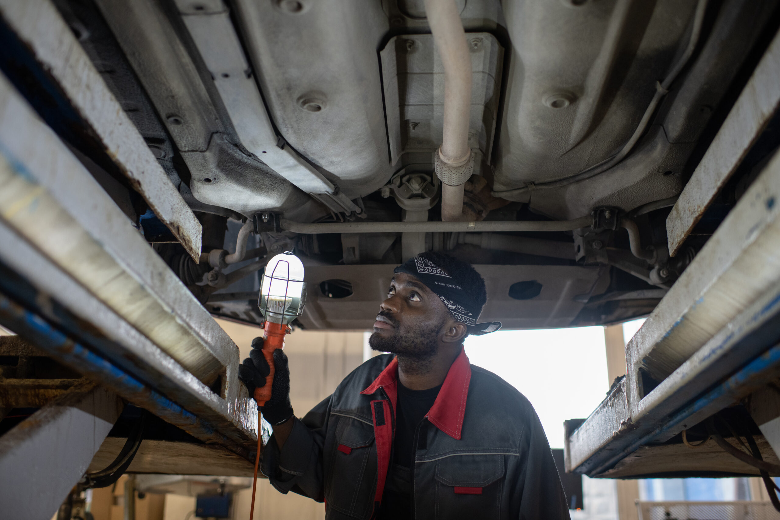 A focused young male auto technician, illuminated by a work lamp, inspects the undercarriage of a car during a repair.