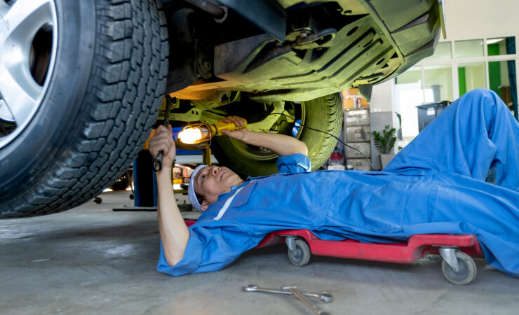 A mechanic in a blue jumpsuit lies on a creeper under a car, using a wrench to tighten a bolt.