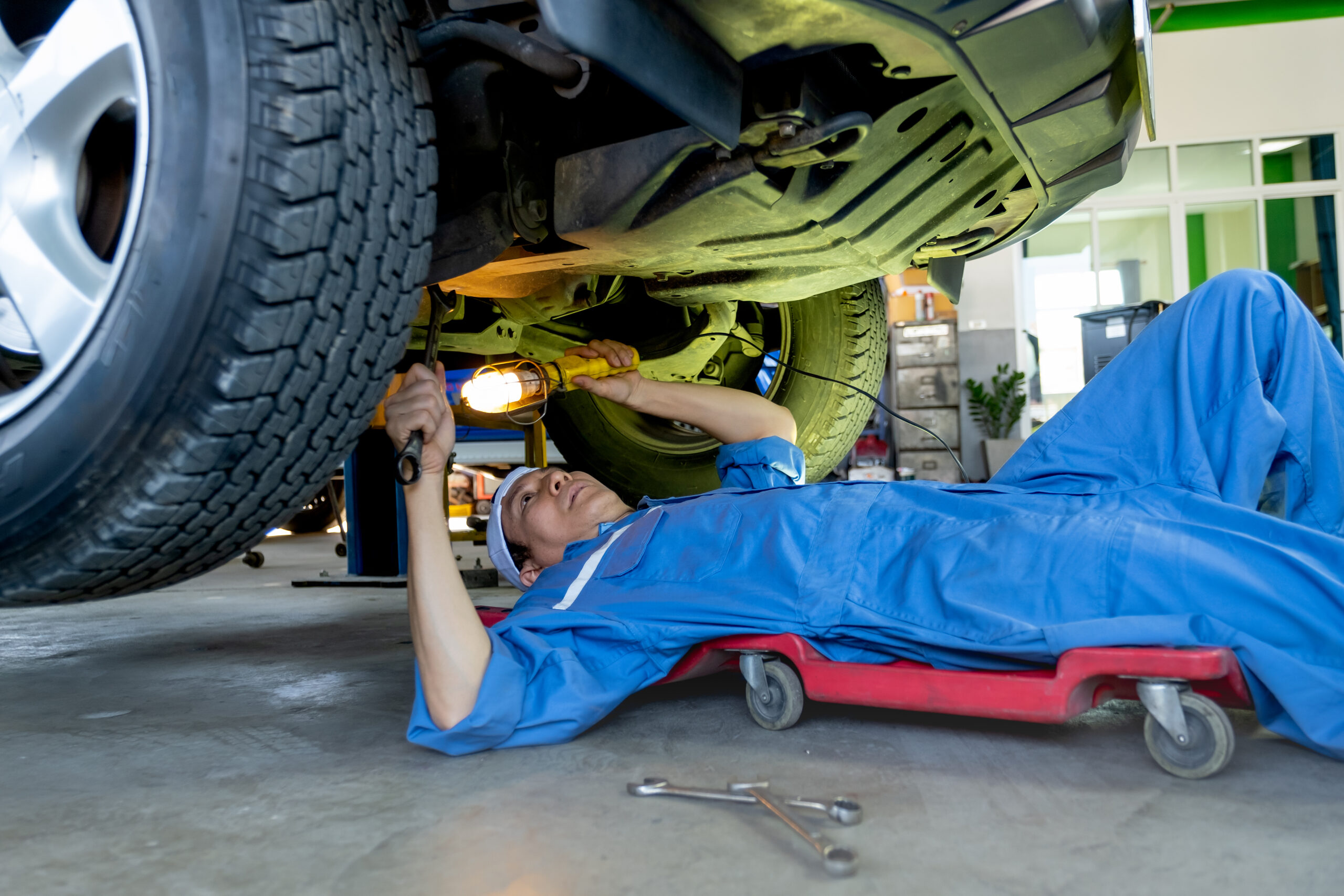 A mechanic in a blue jumpsuit lies on a creeper under a car, using a wrench to tighten a bolt.