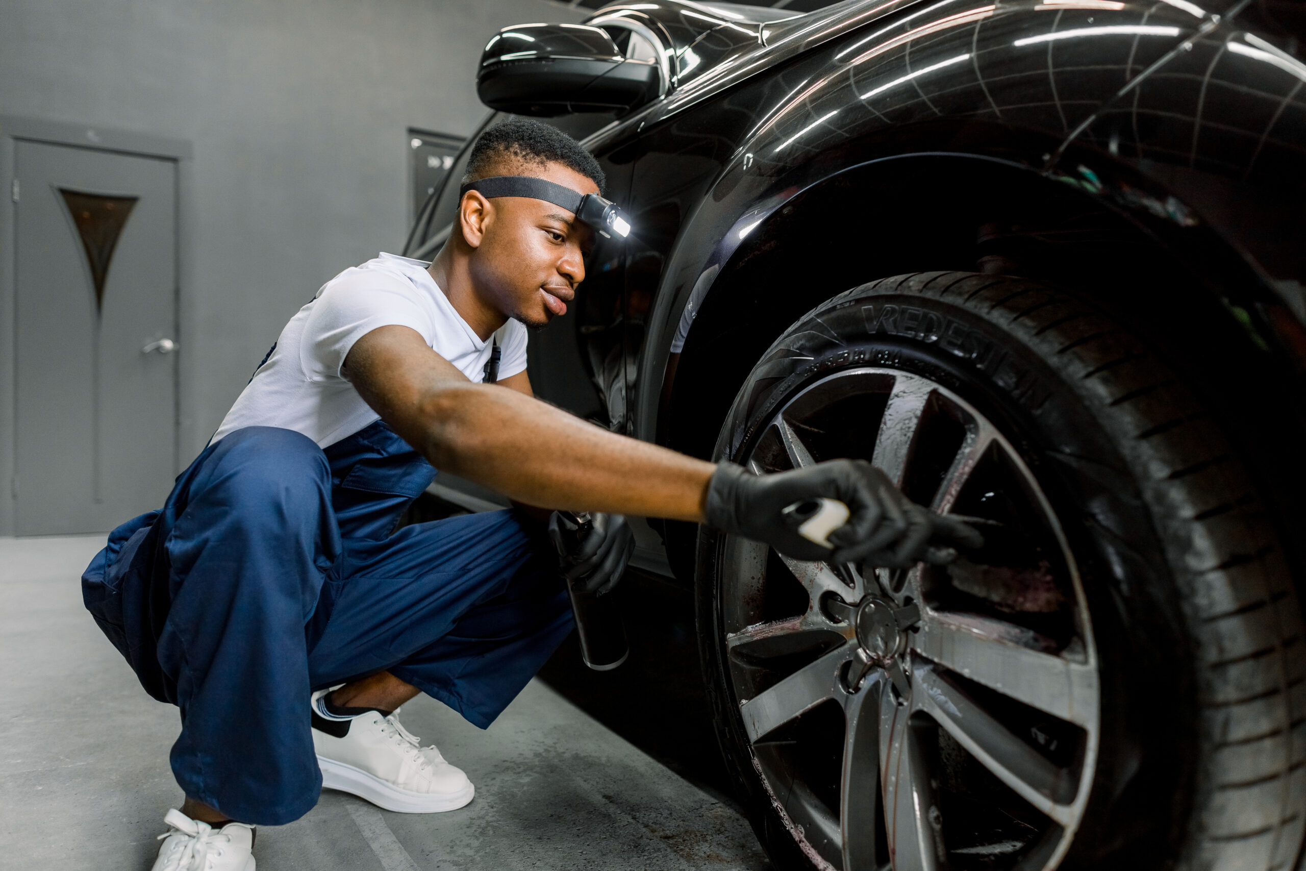 A person wearing a blue t-shirt and jeans meticulously hand washes a white car using a sponge and soapy water.