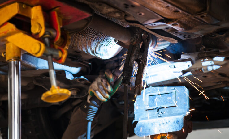 A welder in a protective mask and gloves carefully welds a silencer onto an exhaust pipe, sparks flying.