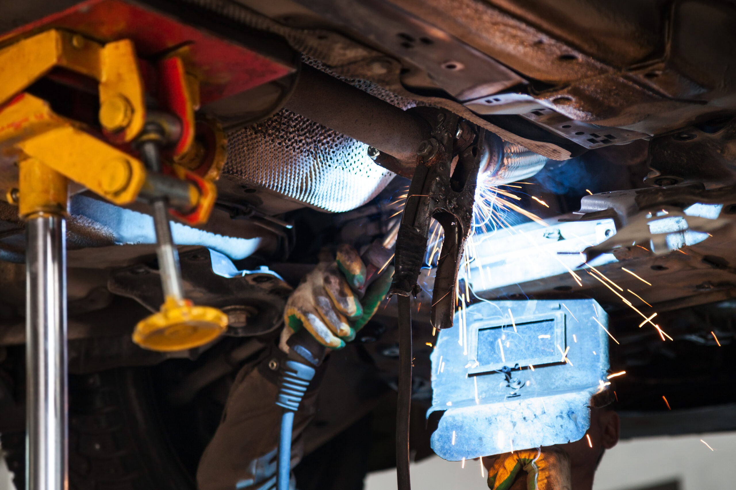 A welder in a protective mask and gloves carefully welds a silencer onto an exhaust pipe, sparks flying.