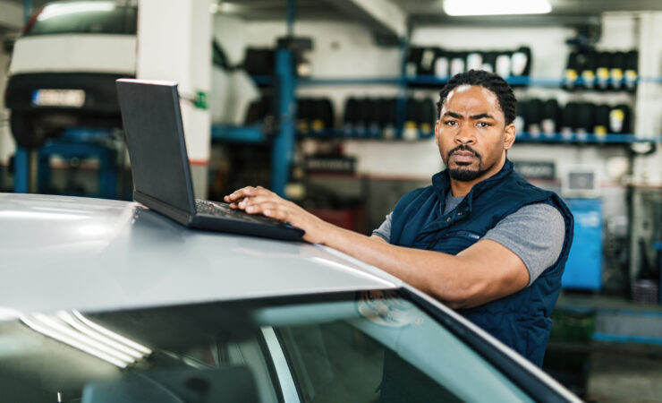 A young Black auto mechanic, wearing a blue jumpsuit and safety glasses, sits at a computer in a modern auto repair shop, analyzing diagnostic data on the screen.