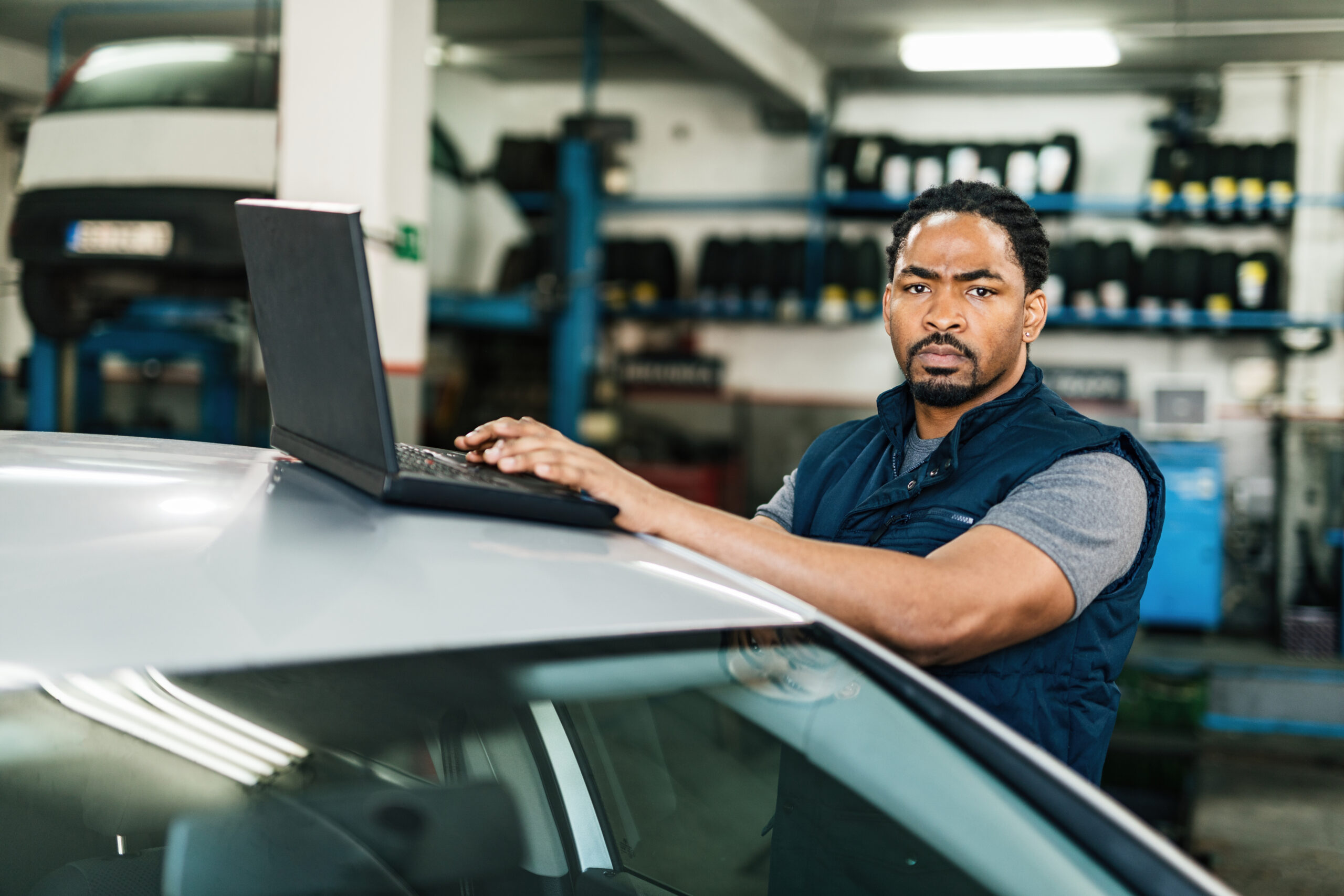 A young Black auto mechanic, wearing a blue jumpsuit and safety glasses, sits at a computer in a modern auto repair shop, analyzing diagnostic data on the screen.