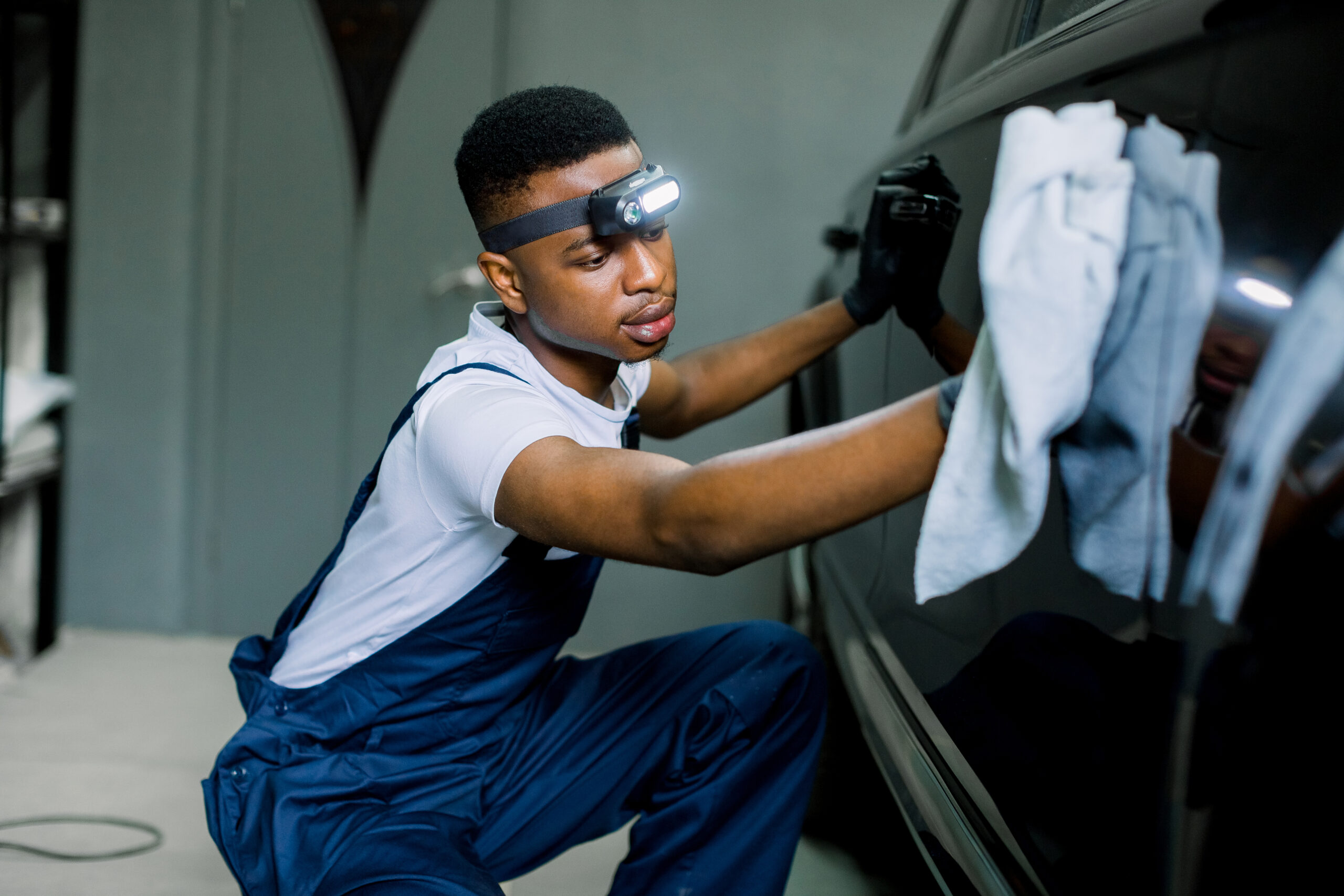 A young Black man in a car wash uniform, smiling as he expertly dries a freshly washed car with a microfiber towel.