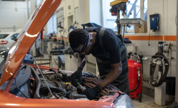 A young Black mechanic, wearing a blue jumpsuit and safety glasses, leans over a car engine, using a wrench to tighten a bolt.c