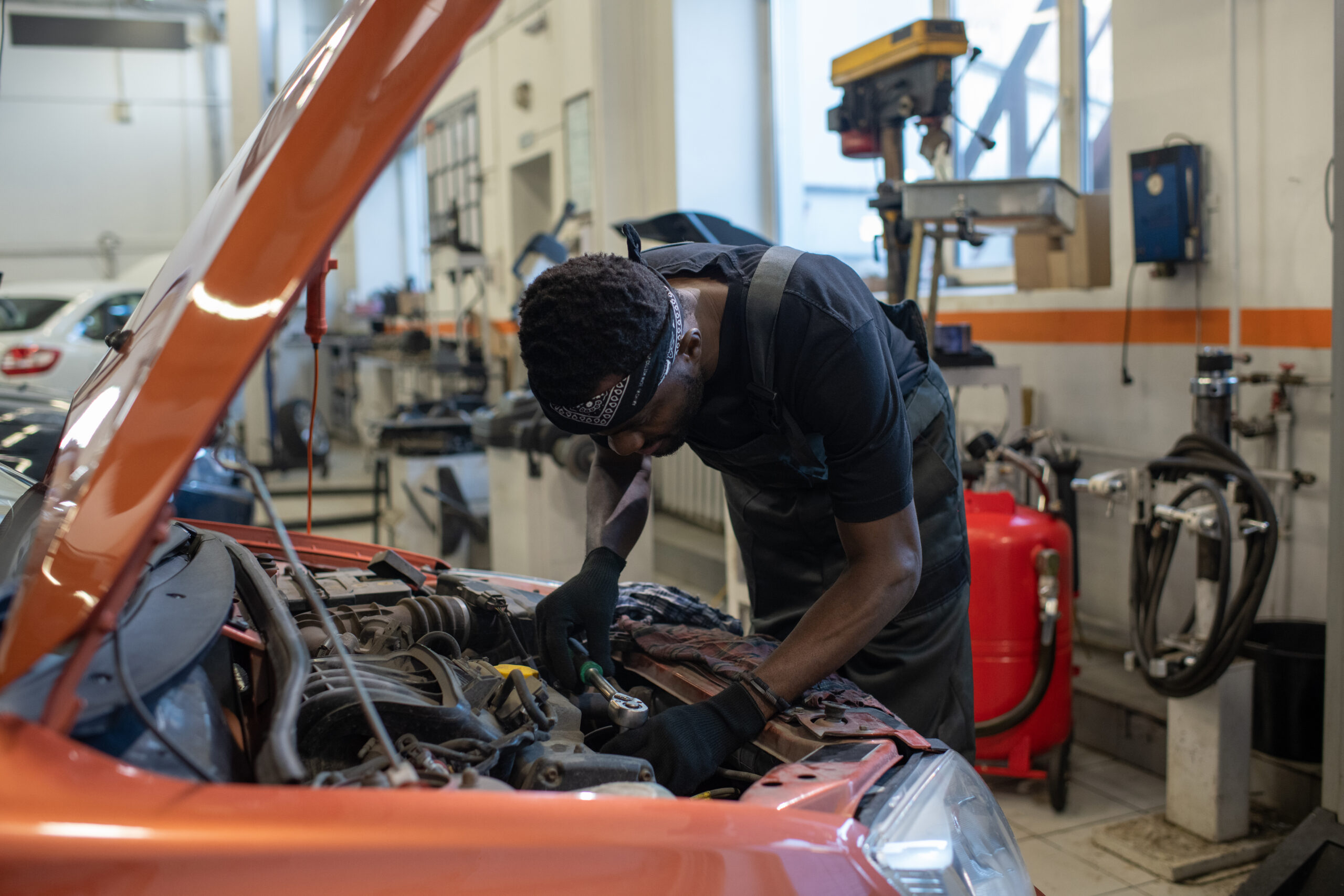 A young Black mechanic, wearing a blue jumpsuit and safety glasses, leans over a car engine, using a wrench to tighten a bolt.c