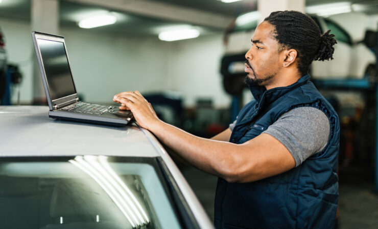 A young Black mechanic, dressed in a blue jumpsuit, sits at a computer in an auto repair shop, carefully reviewing diagnostic data.