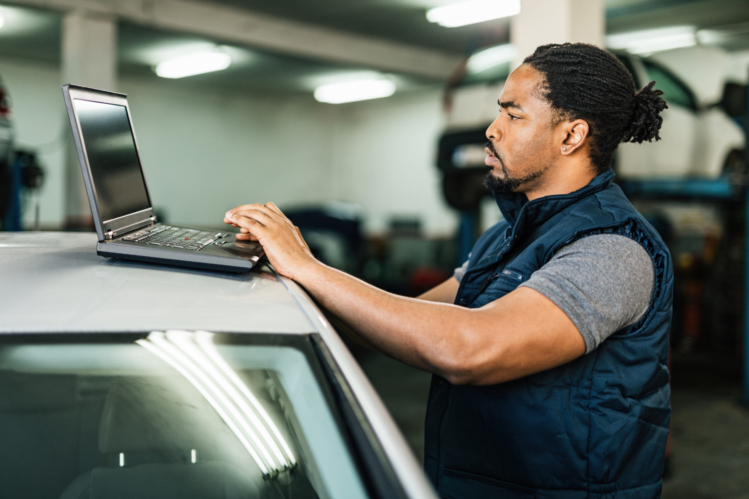 A young Black mechanic, dressed in a blue jumpsuit, sits at a computer in an auto repair shop, carefully reviewing diagnostic data.