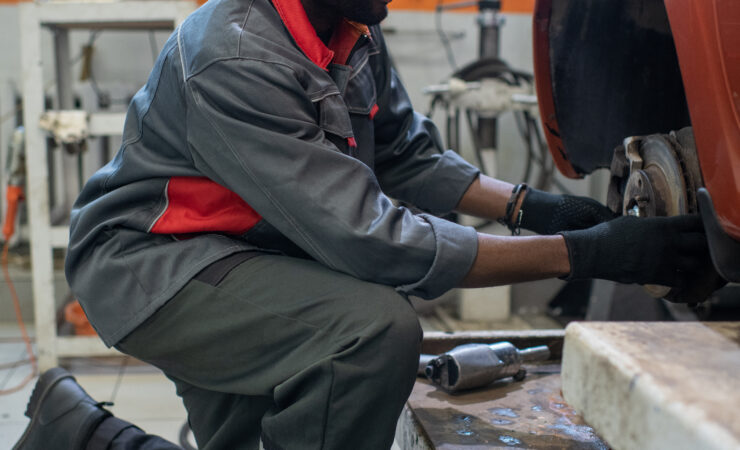 A young mechanic, wearing a blue jumpsuit and work boots, kneels beside a car, using a lug wrench to remove a wheel.