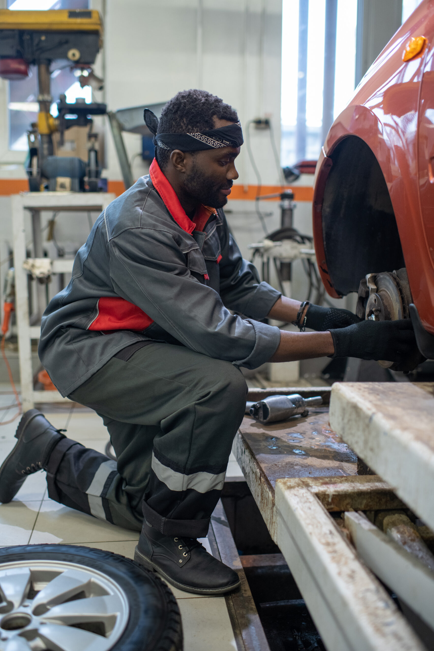 A young mechanic, wearing a blue jumpsuit and work boots, kneels beside a car, using a lug wrench to remove a wheel.