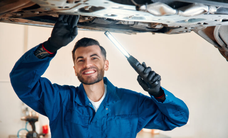 A mechanic in a blue jumpsuit works under the hood of a car in a well-lit garage.