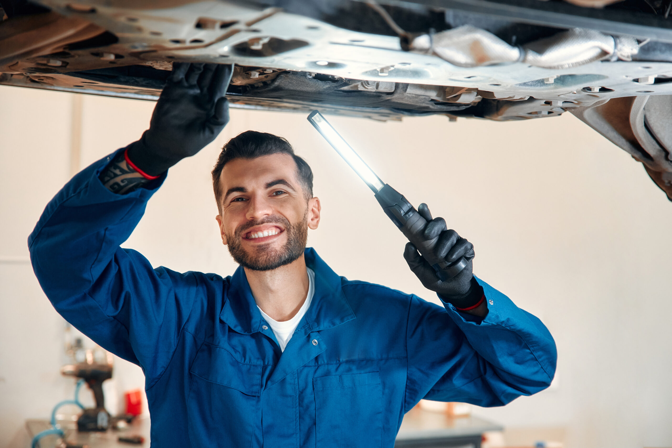 A mechanic in a blue jumpsuit works under the hood of a car in a well-lit garage.