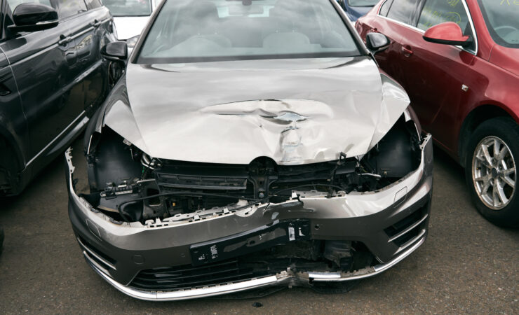 A badly damaged car, with its front end crumpled and missing parts, sits in a car recycling yard.