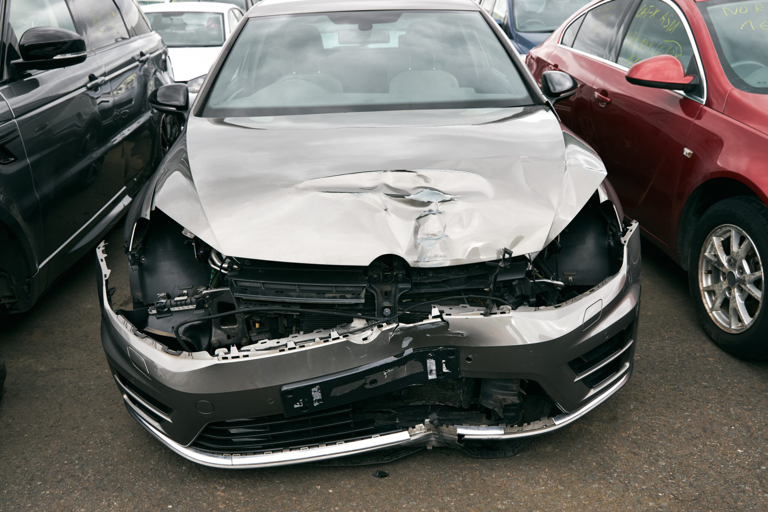 A badly damaged car, with its front end crumpled and missing parts, sits in a car recycling yard.