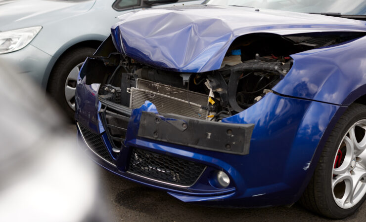 A damaged car with a crumpled hood and shattered windshield sits in a repair shop garage.