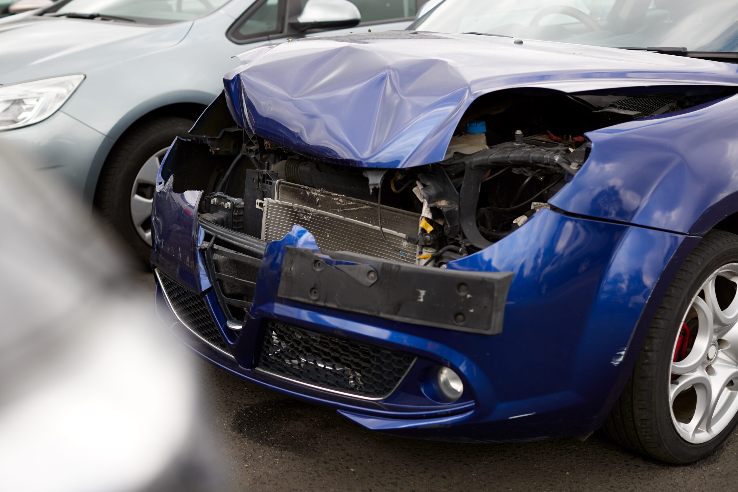 A damaged car with a crumpled hood and shattered windshield sits in a repair shop garage.