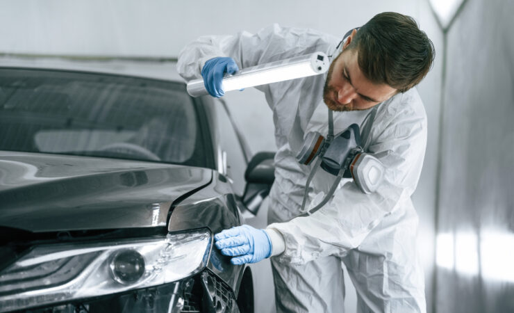 A garage worker, holding a flashlight, carefully inspects the surface of a car for any damage.