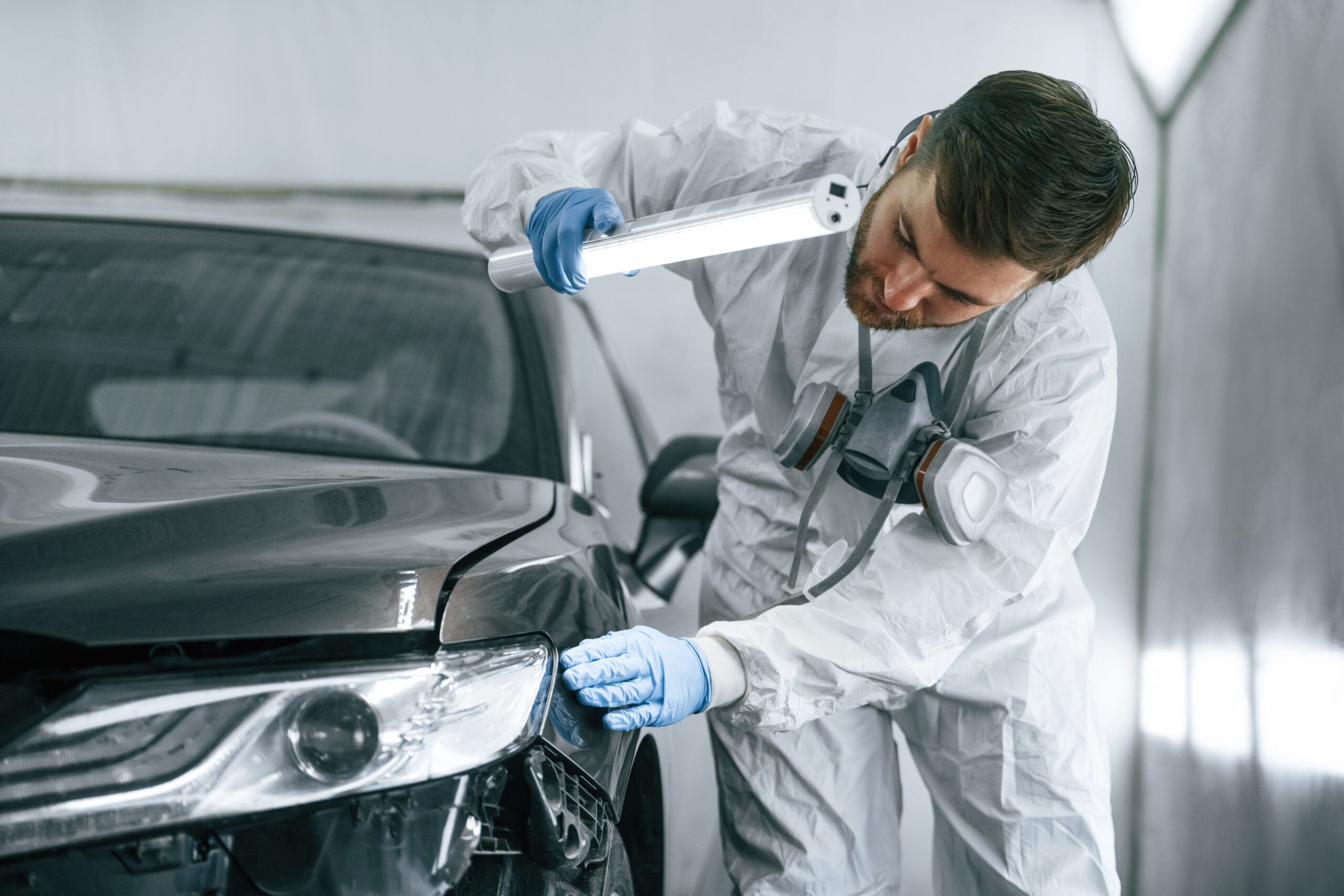 A garage worker, holding a flashlight, carefully inspects the surface of a car for any damage.