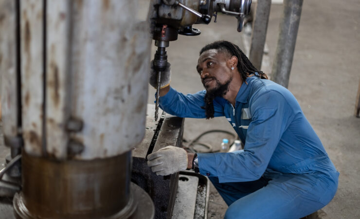 A Black male mechanic, wearing a blue jumpsuit and holding a wrench, inspects a car engine.