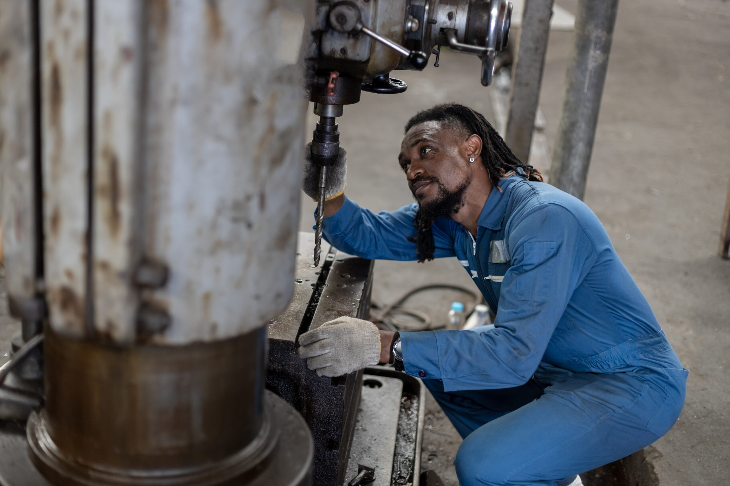A Black male mechanic, wearing a blue jumpsuit and holding a wrench, inspects a car engine.