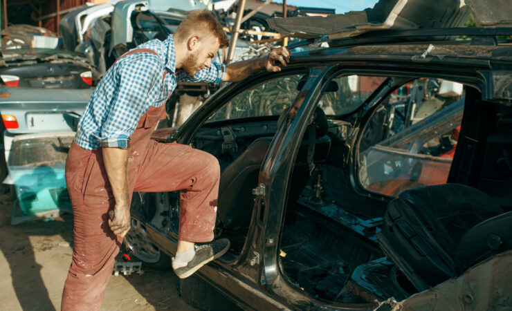 A male mechanic, wearing overalls and work gloves, uses tools to dismantle a car in a junkyard.