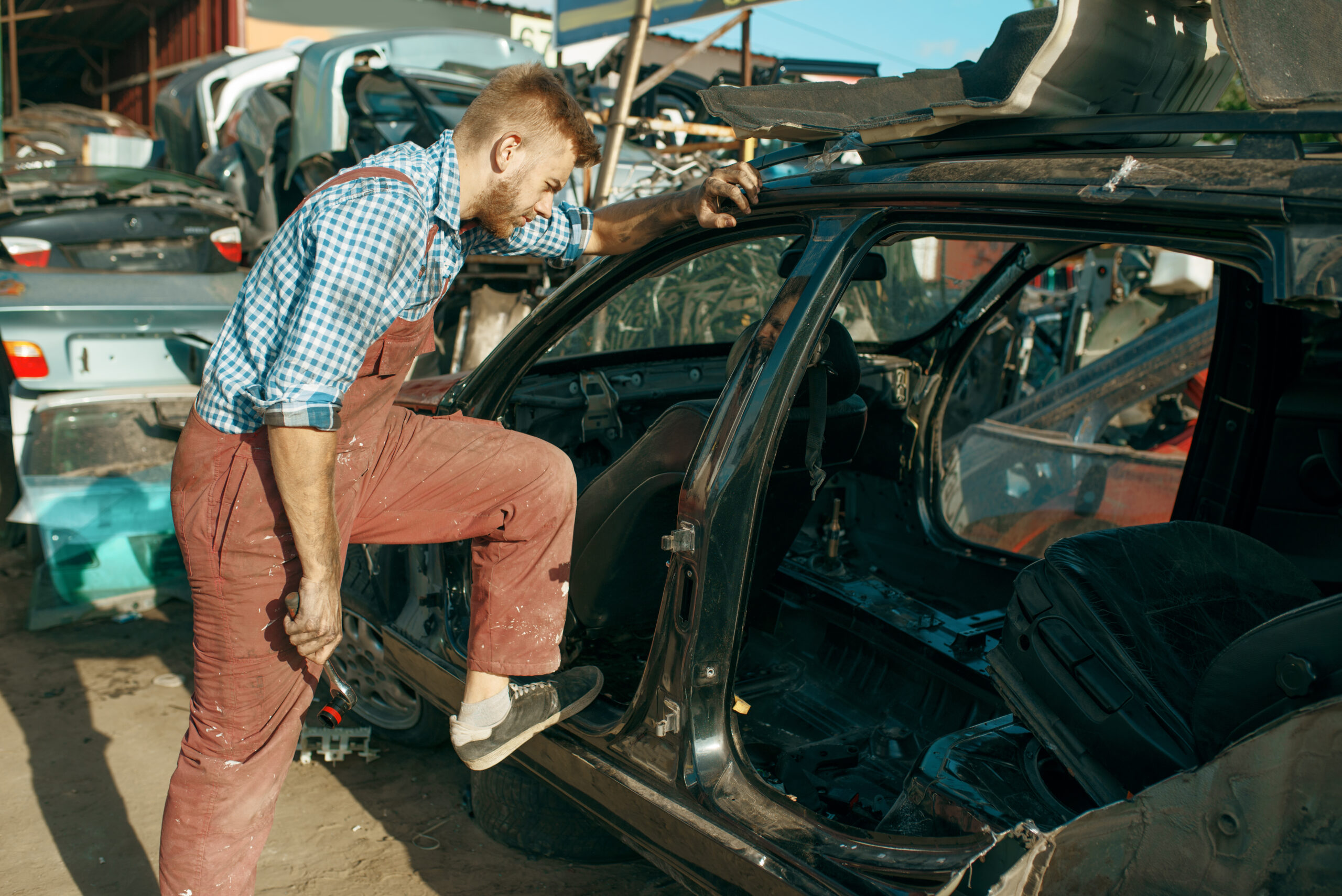 A male mechanic, wearing overalls and work gloves, uses tools to dismantle a car in a junkyard.