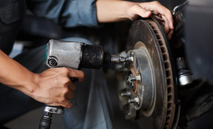 A close-up photo of a mechanic's hands changing a car tire. The mechanic is wearing gloves and using a tire iron to remove the lug nuts. The tire is flat and the rim is dirty.