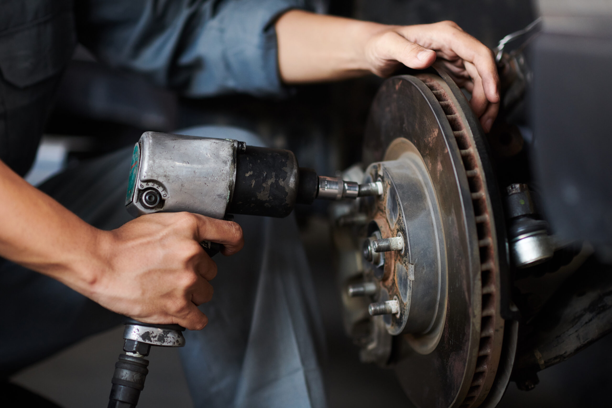 A close-up photo of a mechanic's hands changing a car tire. The mechanic is wearing gloves and using a tire iron to remove the lug nuts. The tire is flat and the rim is dirty.