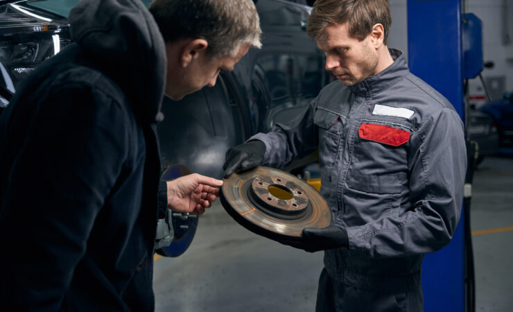 A mechanic, holding a brake disc, discusses car repairs with a client.