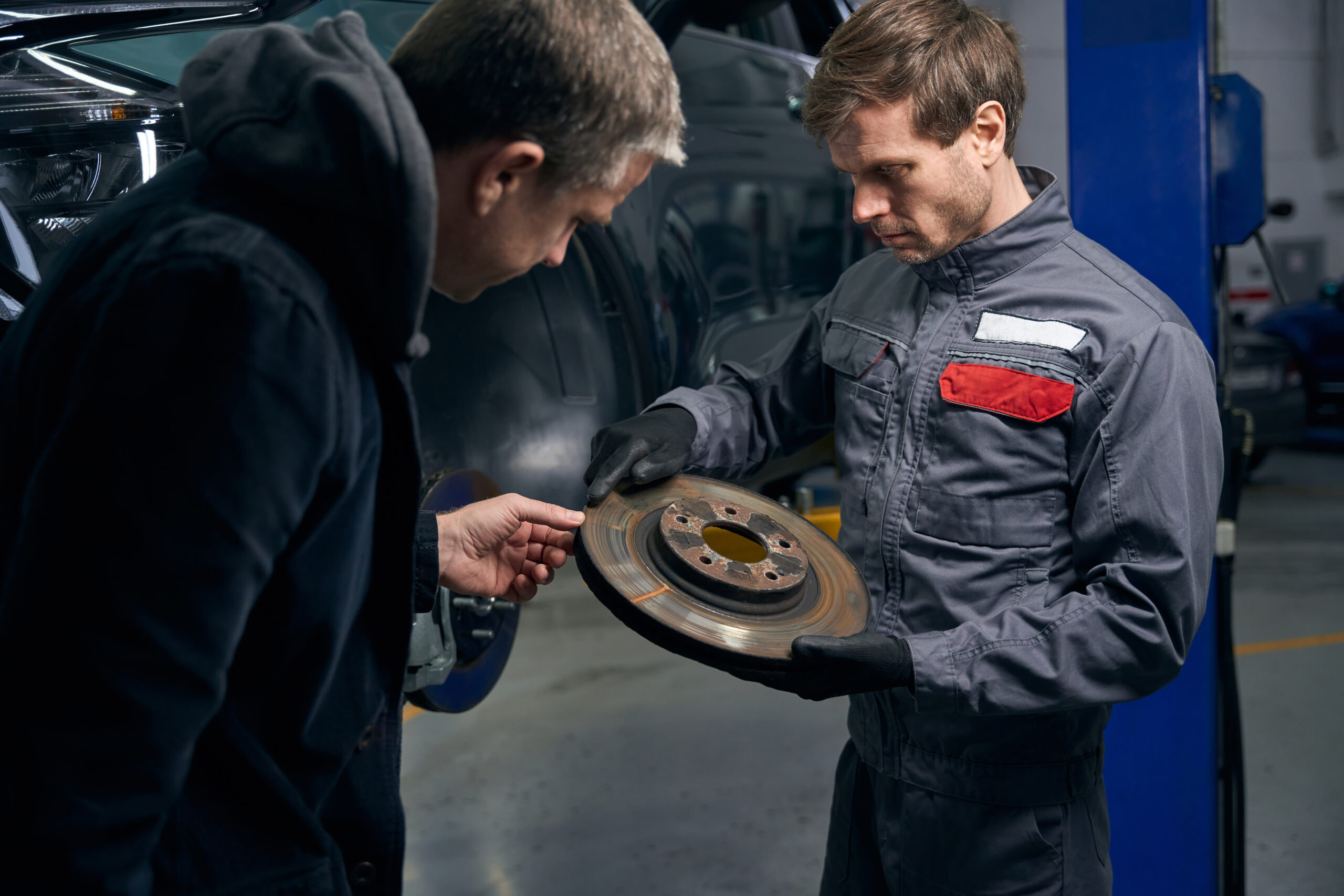 A mechanic, holding a brake disc, discusses car repairs with a client.